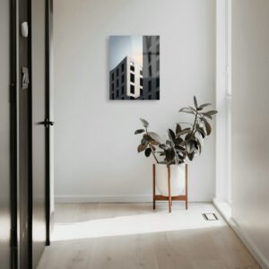 A vertical 14x18 HD metal art print of modern urban architecture mounted on a bright white hallway wall in a contemporary minimalist home, positioned above a potted rubber plant in a wood stand.