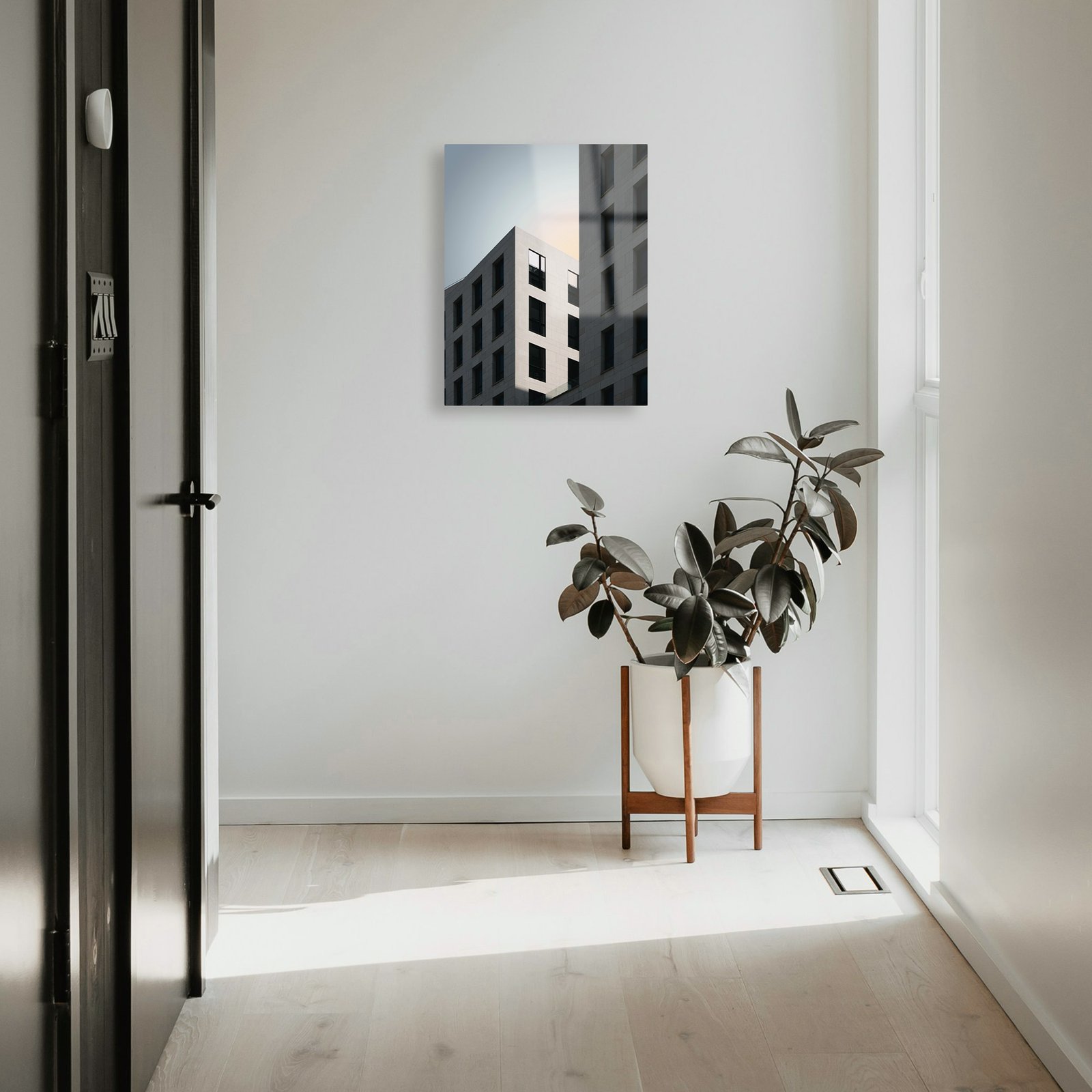 A vertical 14x18 HD metal art print of modern urban architecture mounted on a bright white hallway wall in a contemporary minimalist home, positioned above a potted rubber plant in a wood stand.