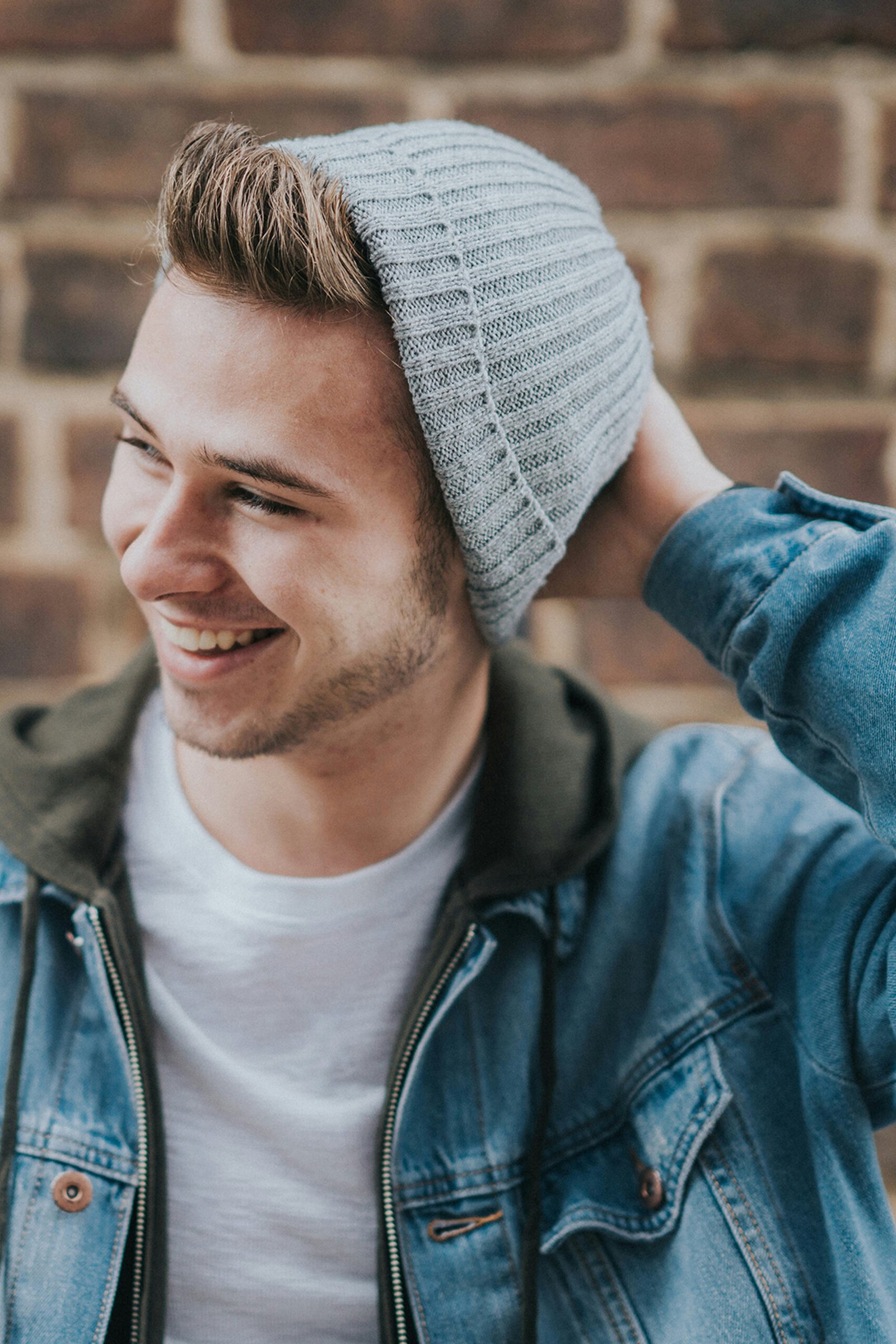 A close-up profile portrait of a smiling young man in his early 20s wearing a grey knit beanie and a denim jacket over a hoodie, standing against a rustic brick wall background.