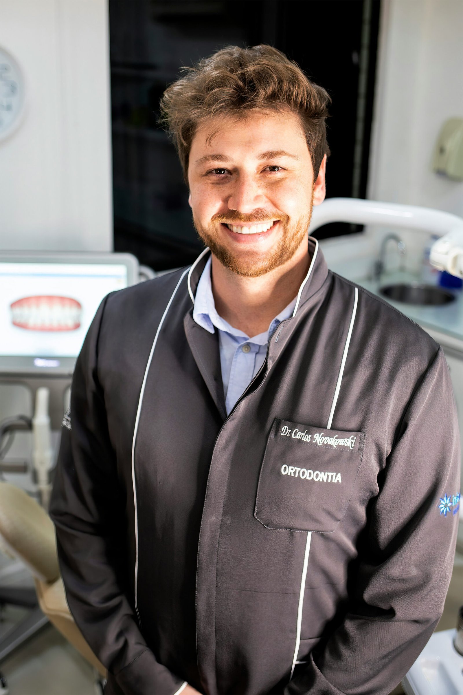 A professional waist-up portrait of a smiling male dentist with brown hair and a beard, wearing a dark grey clinical coat inside a brightly lit modern dental office.