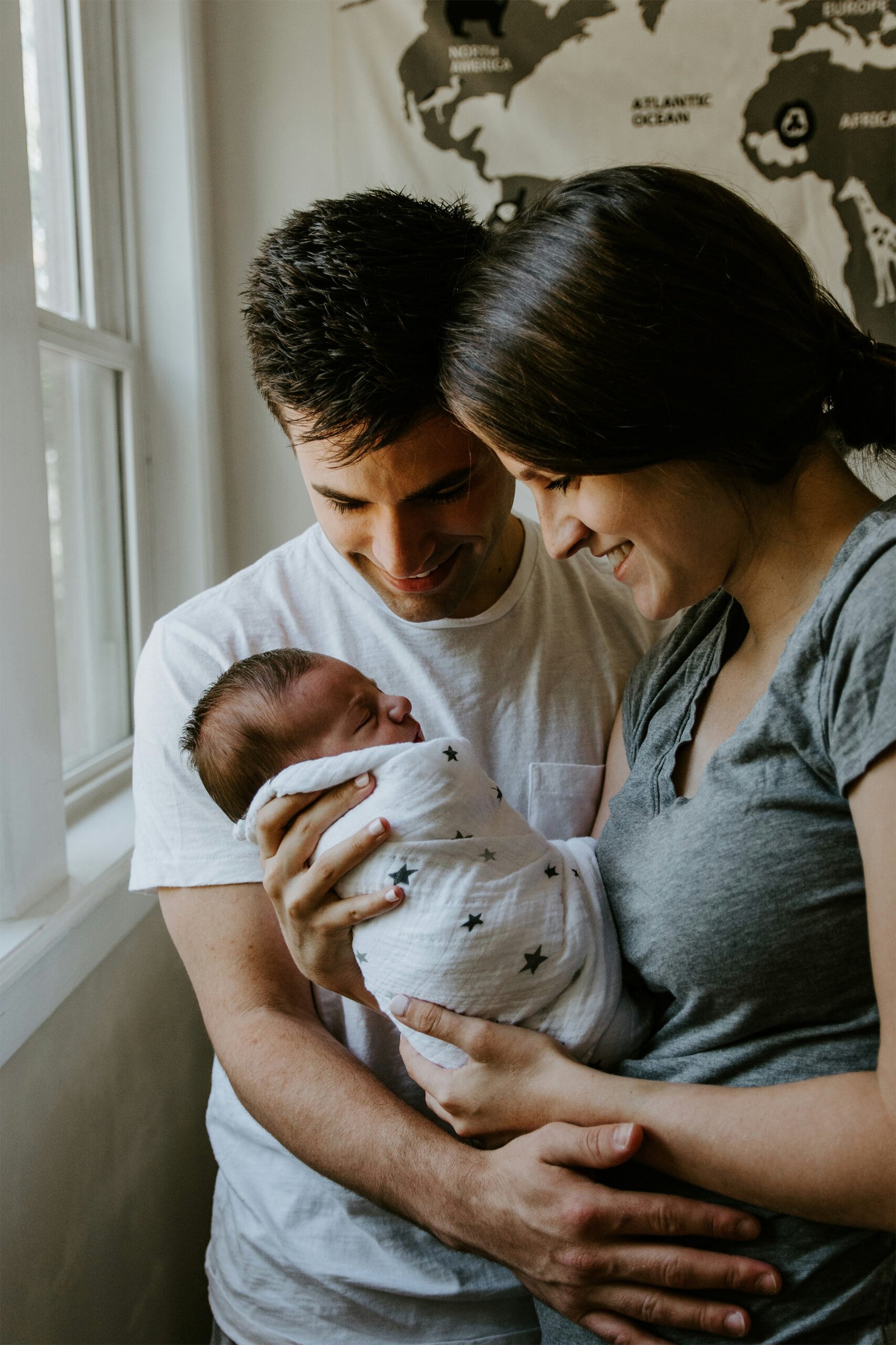 An intimate indoor lifestyle portrait of a mother and father standing together by a bright window, holding and hugging their three young children as warm sunlight streams in from behind.