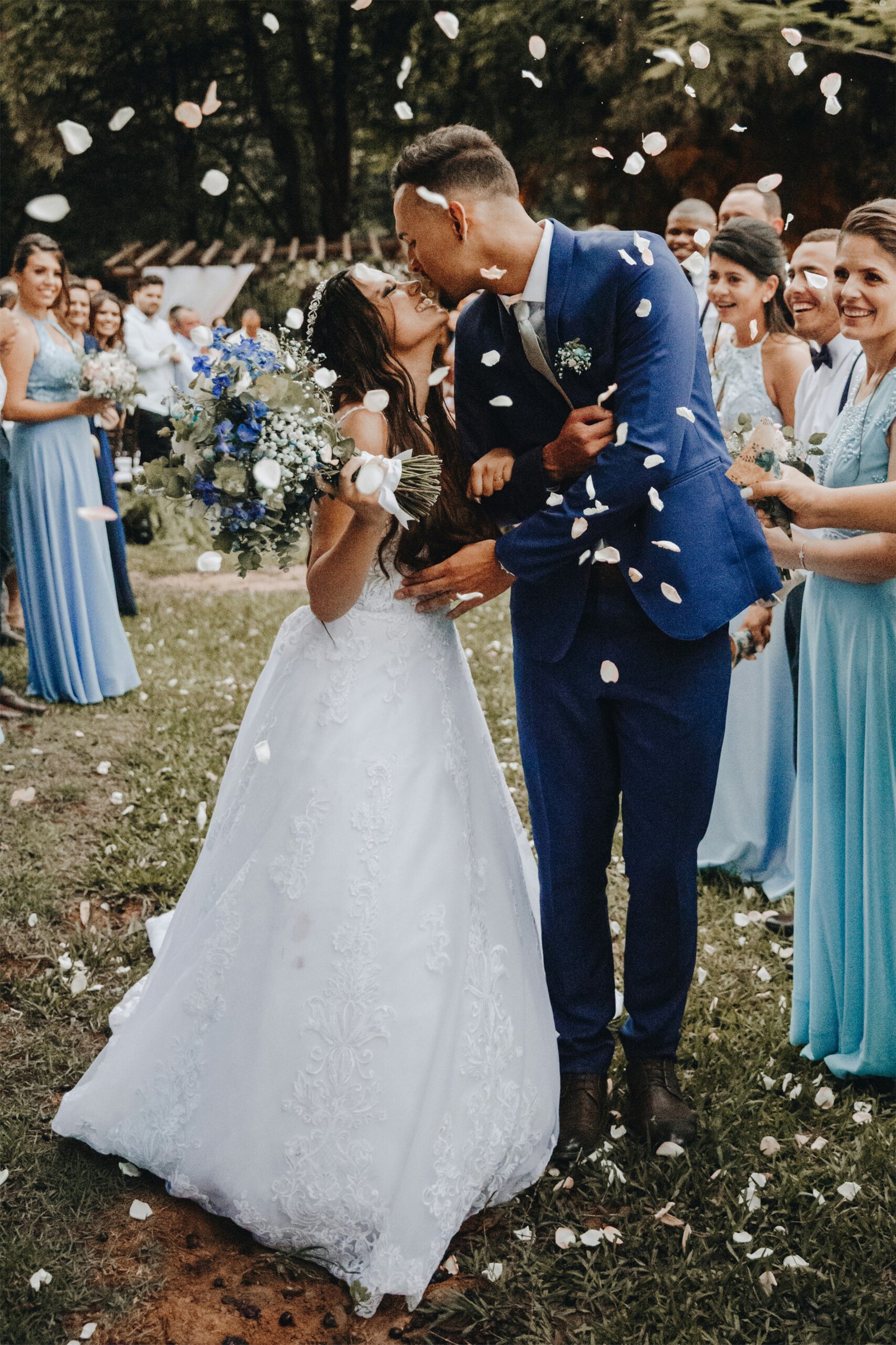 A joyful outdoor wedding lifestyle portrait of a bride in a white lace dress and a groom in a blue suit kissing as guests toss white petals over them.