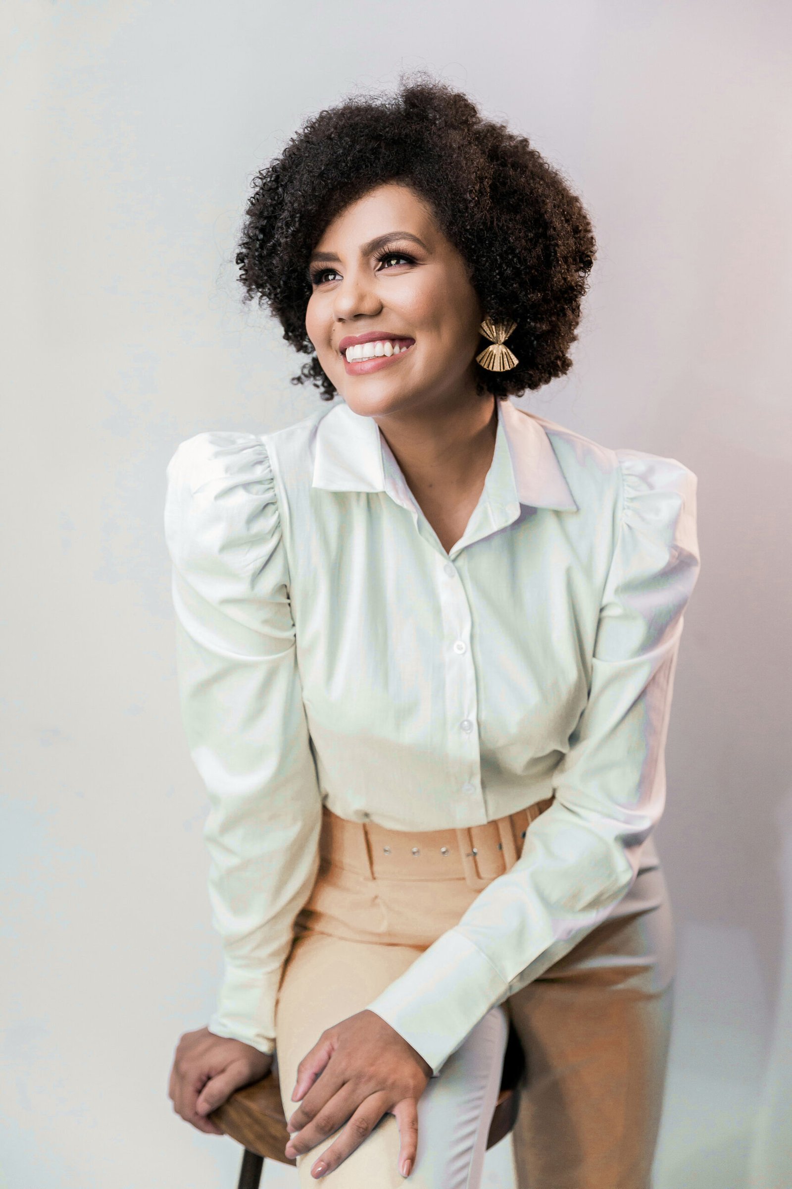 Professional studio portrait of a smiling woman with curly Afro-textured hair, wearing a white puff-sleeve button-down shirt, tan trousers, and gold statement earrings, looking off-camera.