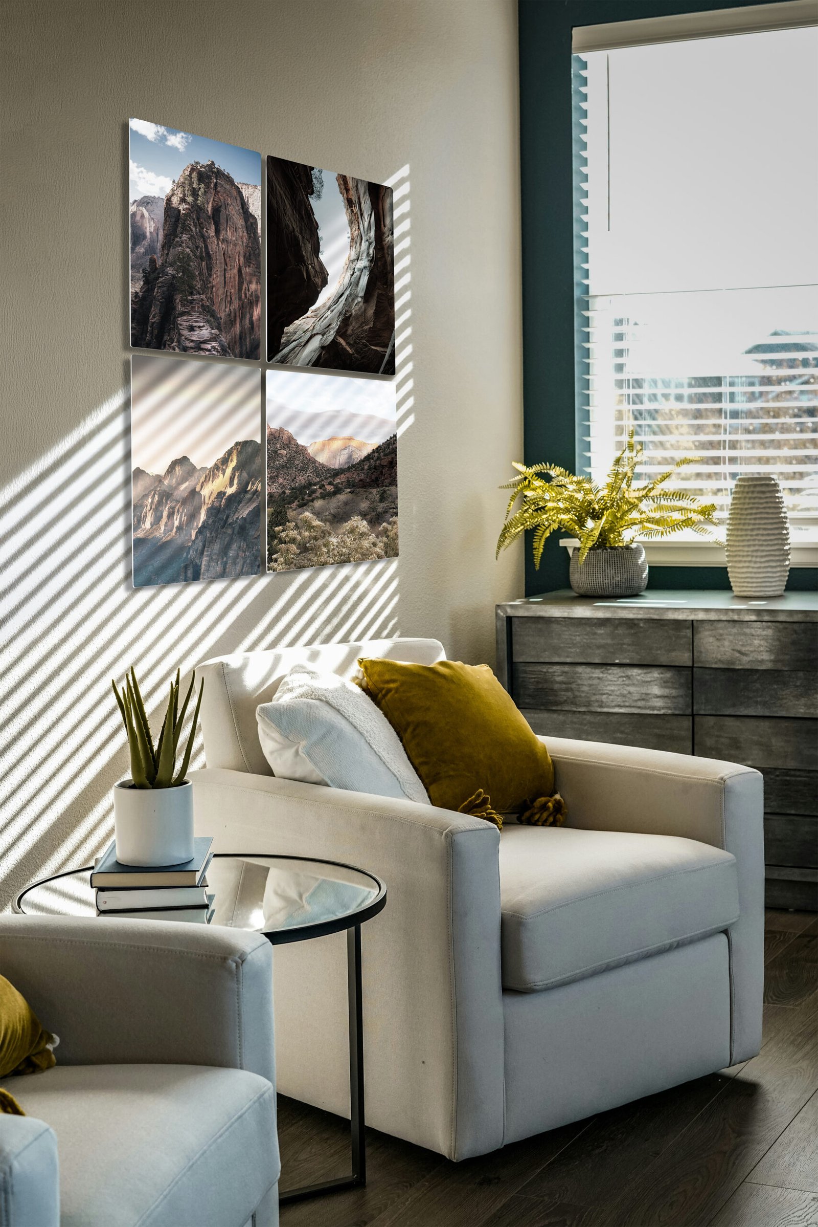 A modern, sunlit therapist's office or waiting room featuring a cozy cream-colored armchair with a mustard yellow throw pillow. On the wall above the chair, a quad-panel grid display showcases four high-gloss square photographs of desert canyon landscapes and mountains, catching the dramatic striped shadows of window blinds. A small glass side table holds a potted succulent and books in the foreground, while a dark wooden dresser with a potted fern sits by the window in the background.