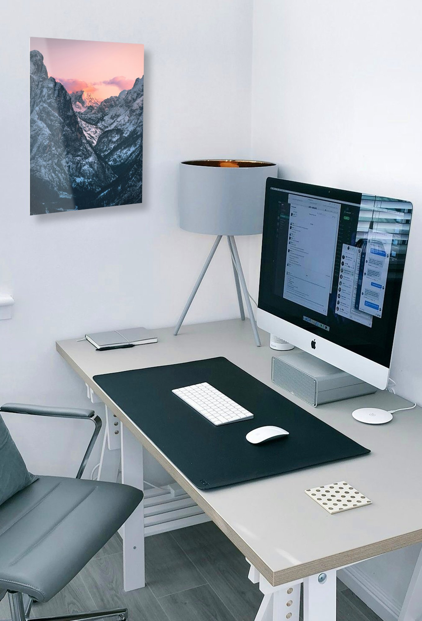 A modern home office desk mockup featuring a single 12x18 vertical mountain landscape metal print mounted on the wall above an iMac and a grey desk lamp.