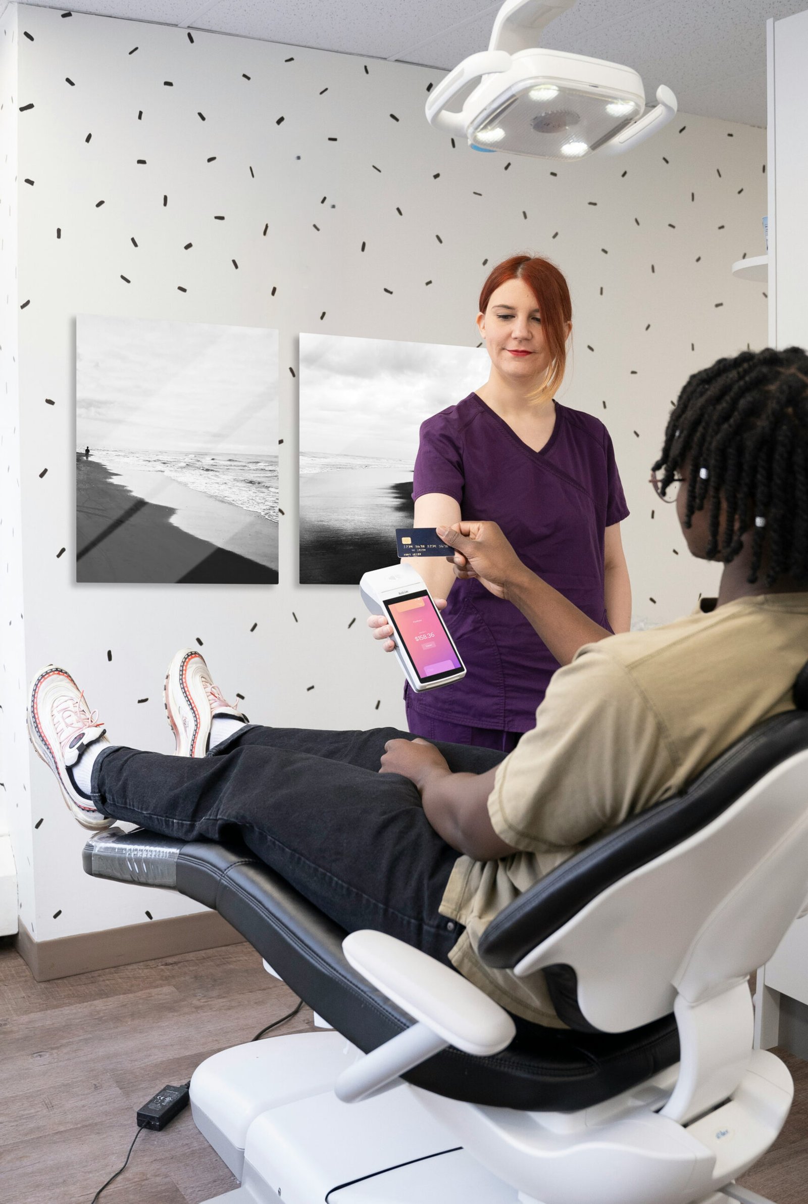 A modern dental clinic check-out mockup featuring a pair of sleek 12x18 vertical black-and-white landscape metal prints mounted side-by-side on a patterned wall above a patient paying in a dental chair.