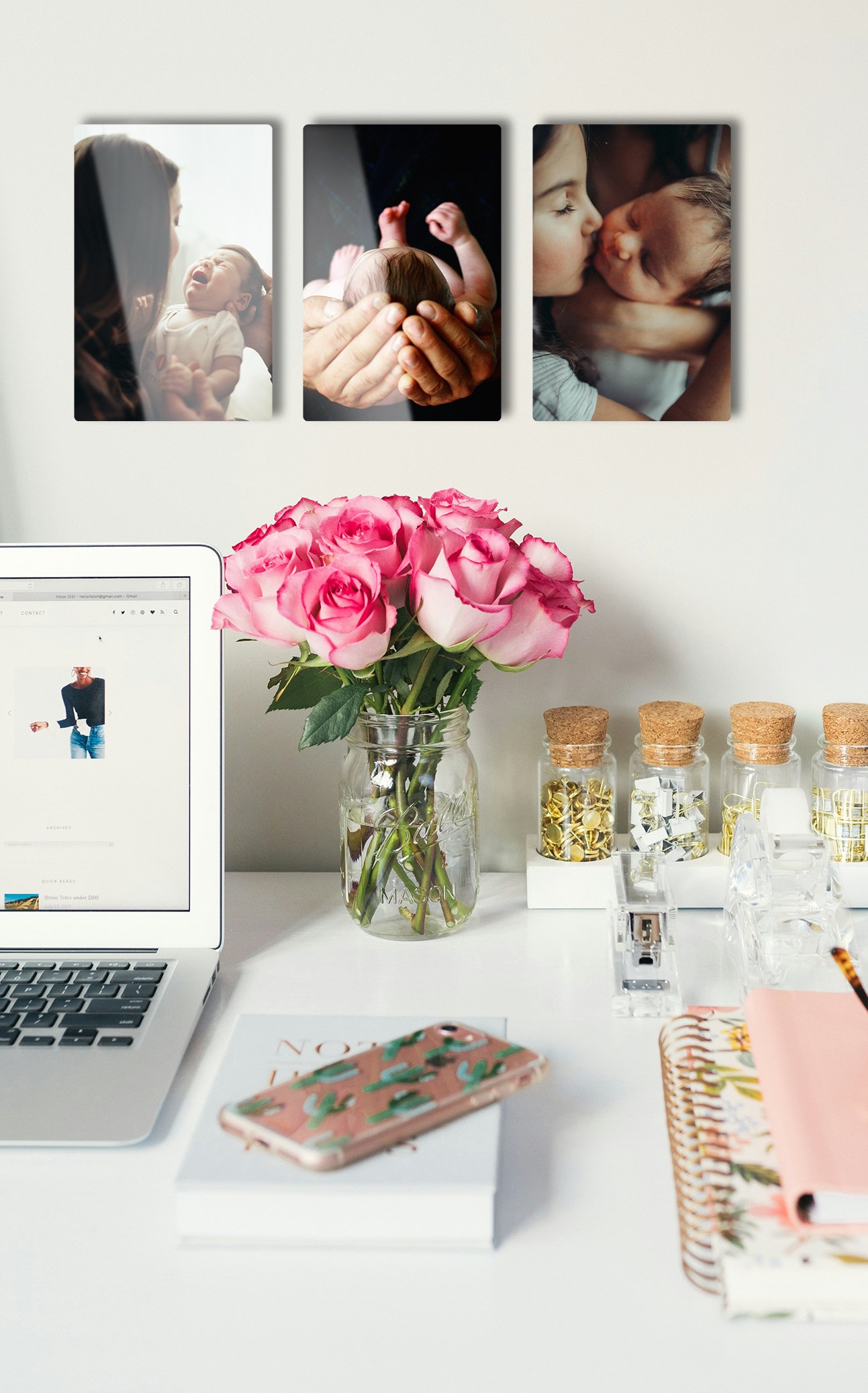 A modern home office desk mockup featuring a vertical 3-panel triptych display of custom 4x6 newborn baby photo metal prints mounted on the wall above a laptop and a vase of pink roses.