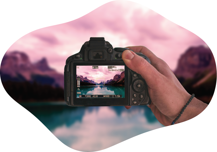 A photographer holding a DSLR camera, framing a landscape photo of mountains reflecting on a lake at sunset.