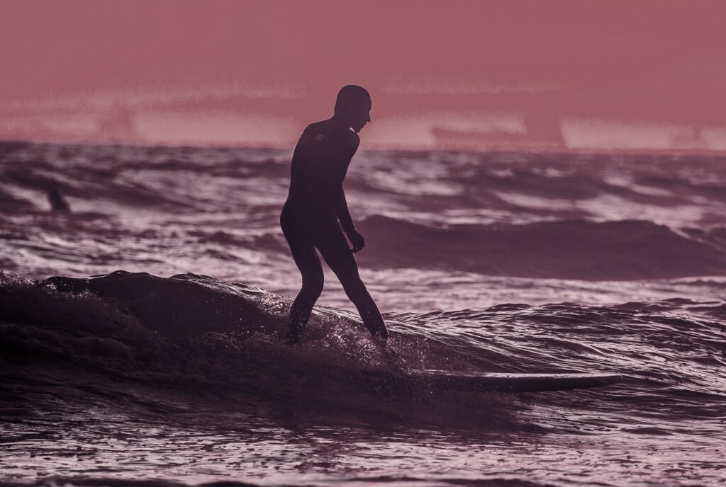 Silhouetted surfer riding an ocean wave in a full wetsuit under a moody pink and purple sunset skyAction sports photograph featuring a surfer silhouetted against dynamic ocean waves at dusk. Toned in muted purple and dusty pink hues, this image is optimized for use as a hero section background, category banner, or full-width promotional graphic for sports, lifestyle, and custom print showcases.
