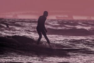 Silhouetted surfer riding an ocean wave in a full wetsuit under a moody pink and purple sunset skyAction sports photograph featuring a surfer silhouetted against dynamic ocean waves at dusk. Toned in muted purple and dusty pink hues, this image is optimized for use as a hero section background, category banner, or full-width promotional graphic for sports, lifestyle, and custom print showcases.