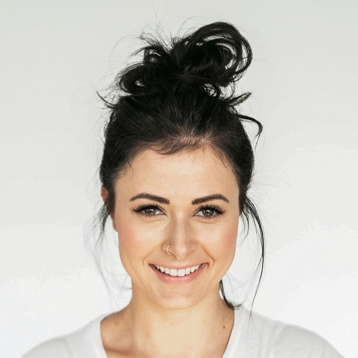Studio portrait of a smiling woman with dark hair in a messy top bun, wearing a white shirt against a plain white background