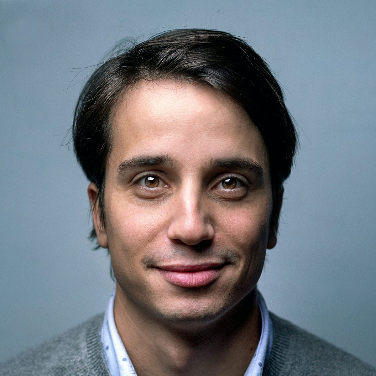 Studio portrait headshot of a man with dark hair, dark eyes, and a slight smile against a solid blue background