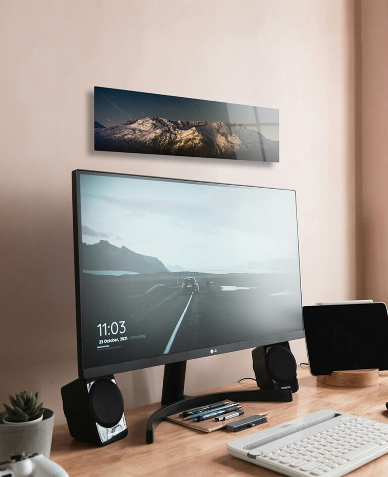 Panoramic metal photo print of a sunset mountain range mounted above a modern desktop computer on a wooden office desk